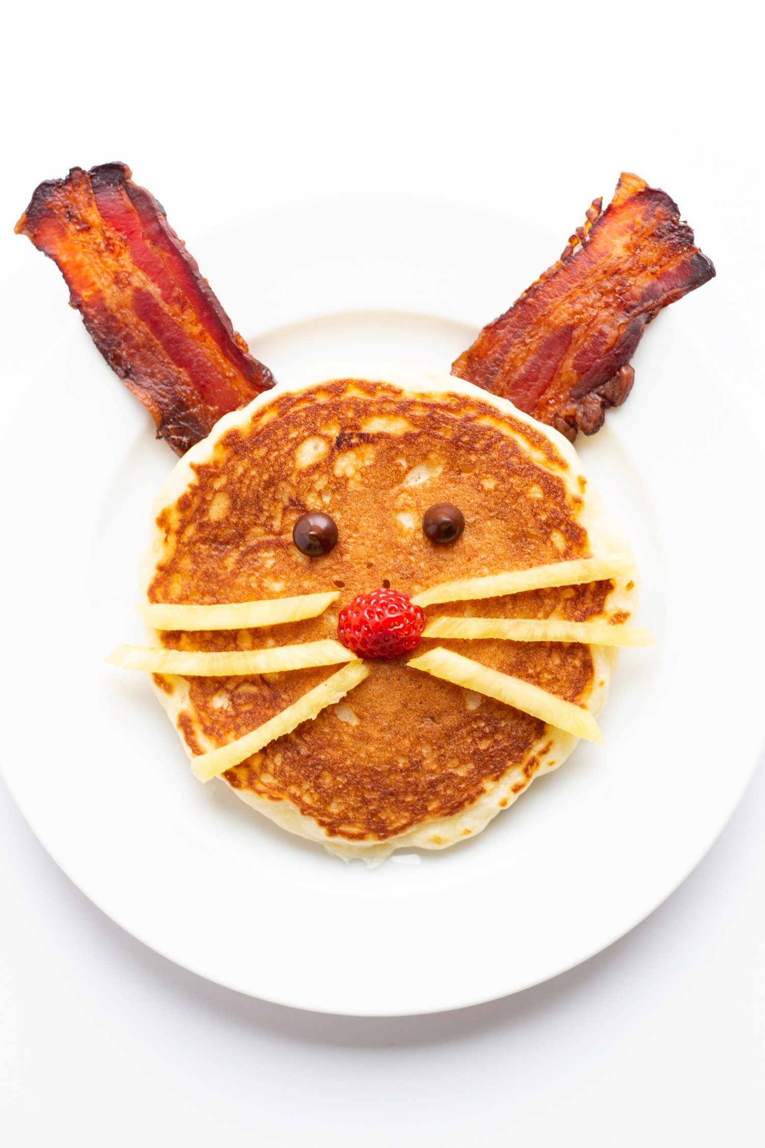 Easter Bunny pancake on a white plate with bacon for ears, chocolate chips for eyes, strawberry for a nose and thin pineapple strips for whiskers.