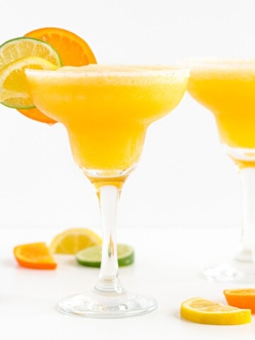 Two margarita glasses filled with frozen citrus margarita and garnished with orange, lemon and lime slices.