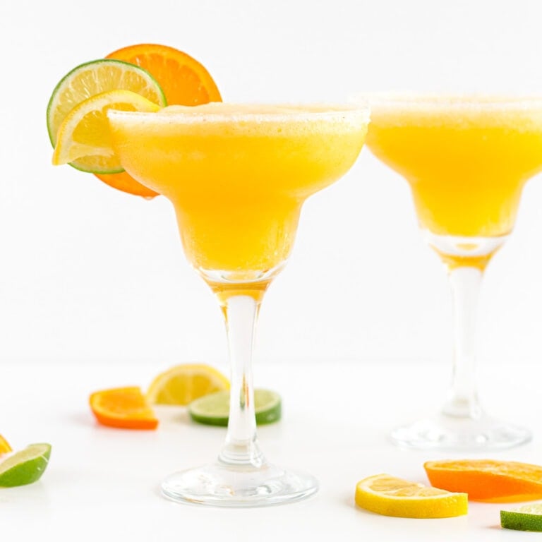 Two margarita glasses filled with frozen citrus margarita and garnished with orange, lemon and lime slices.