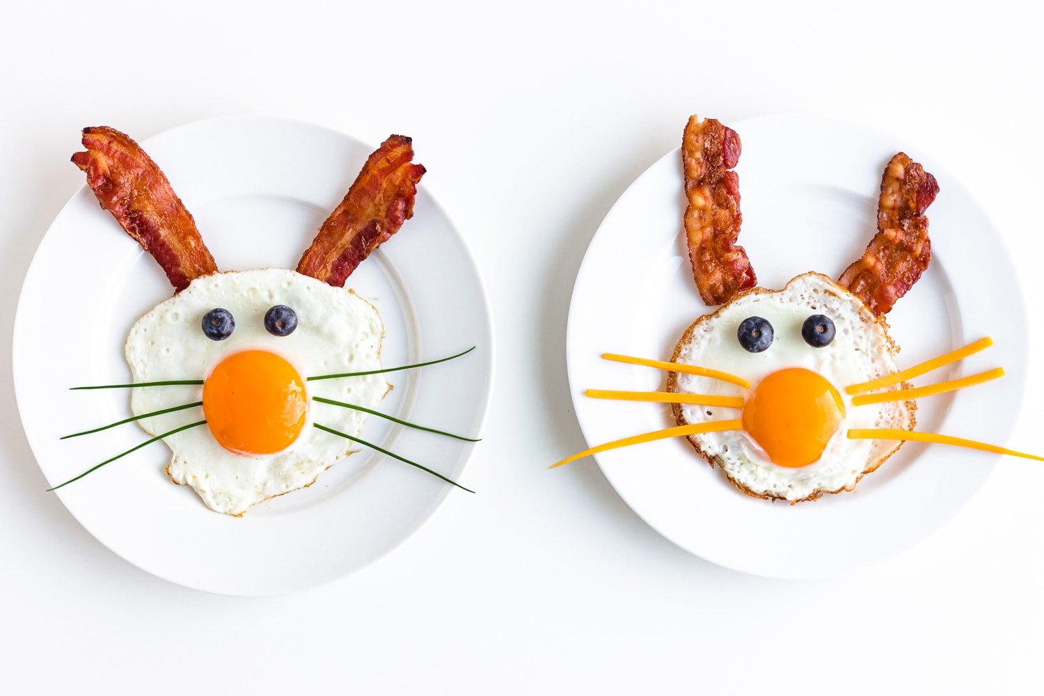 Two fried egg bunnies on white plates with bacon for ears, blueberries for eyes and chives or cheese for whiskers.