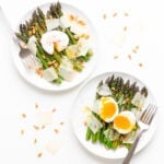 Two plates of roasted asparagus salad, one topped with a poached egg and the other topped with a soft boiled egg.