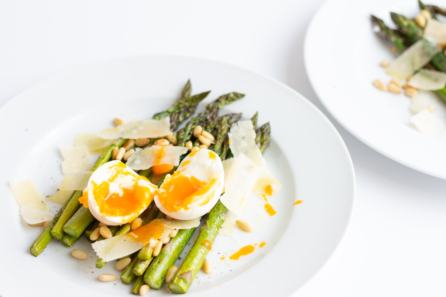 roasted asparagus on a white plate topped with soft boiled egg, parmesan shavings and toasted pine nuts