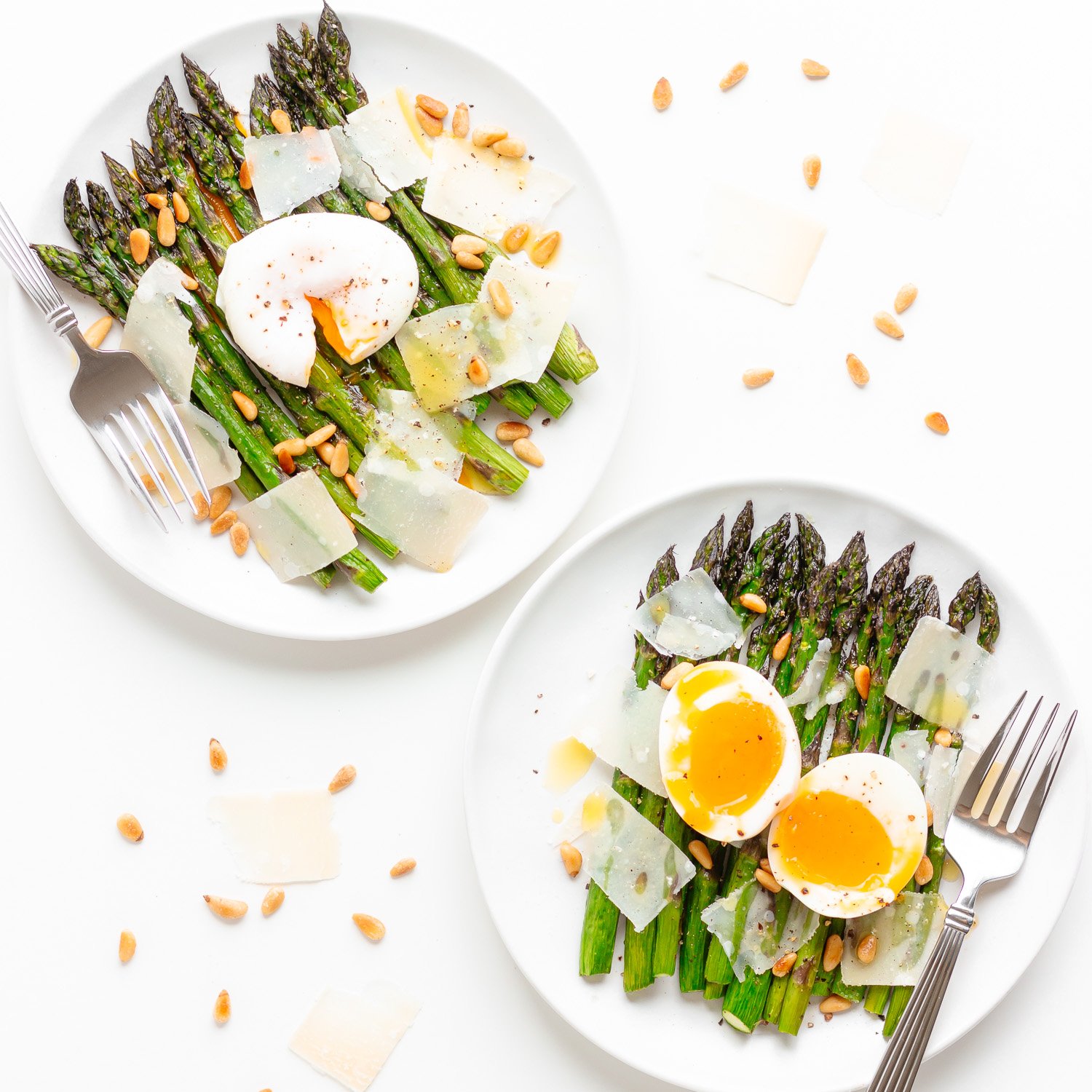 Two plates of roasted asparagus salad topped with egg (one a poached egg and the other a soft boiled egg), parmesan shavings and pine nuts.