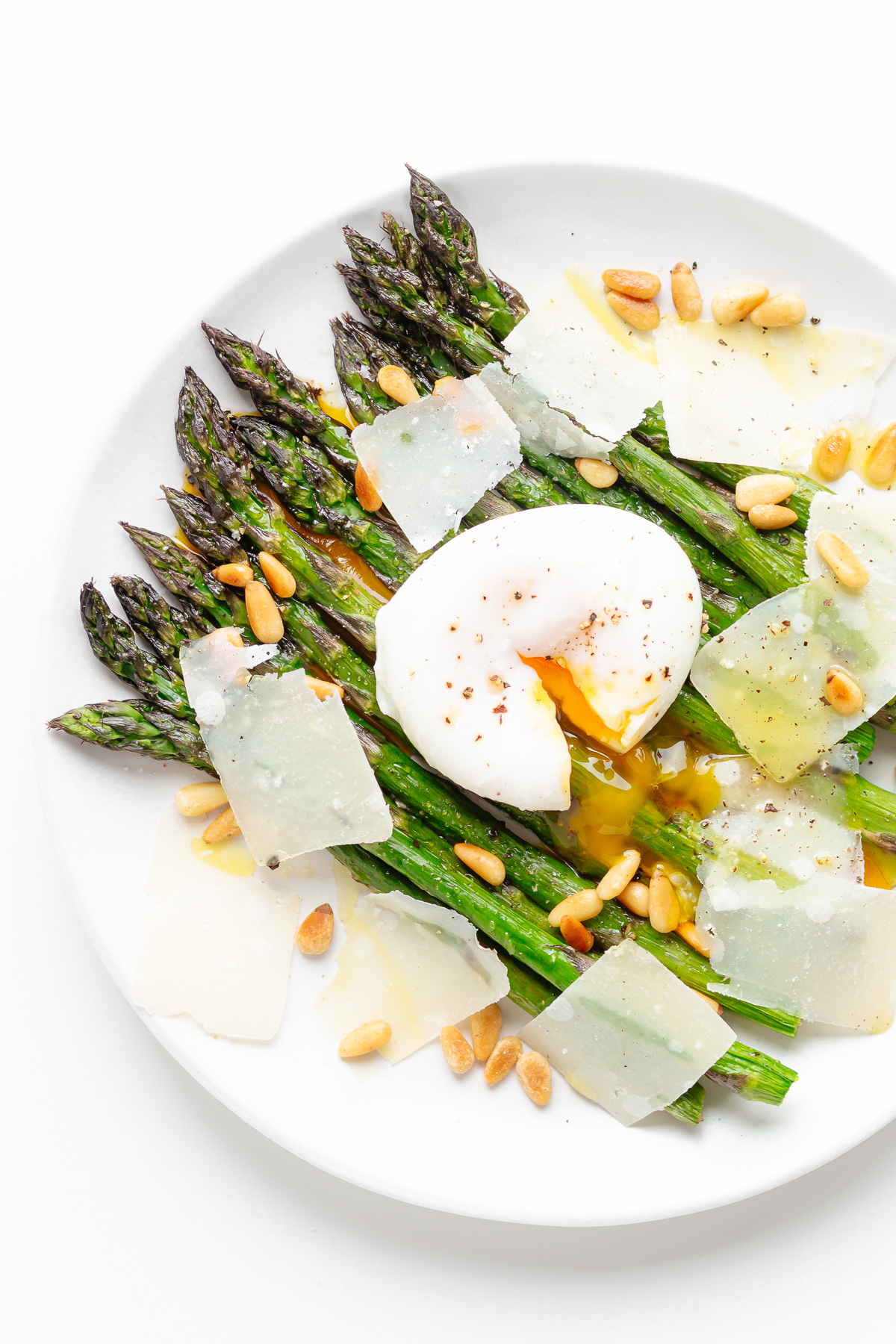 Roasted asparagus on a white plate topped with a poached egg, parmesan shavings, pine nuts, drizzle of olive oil and salt and pepper.