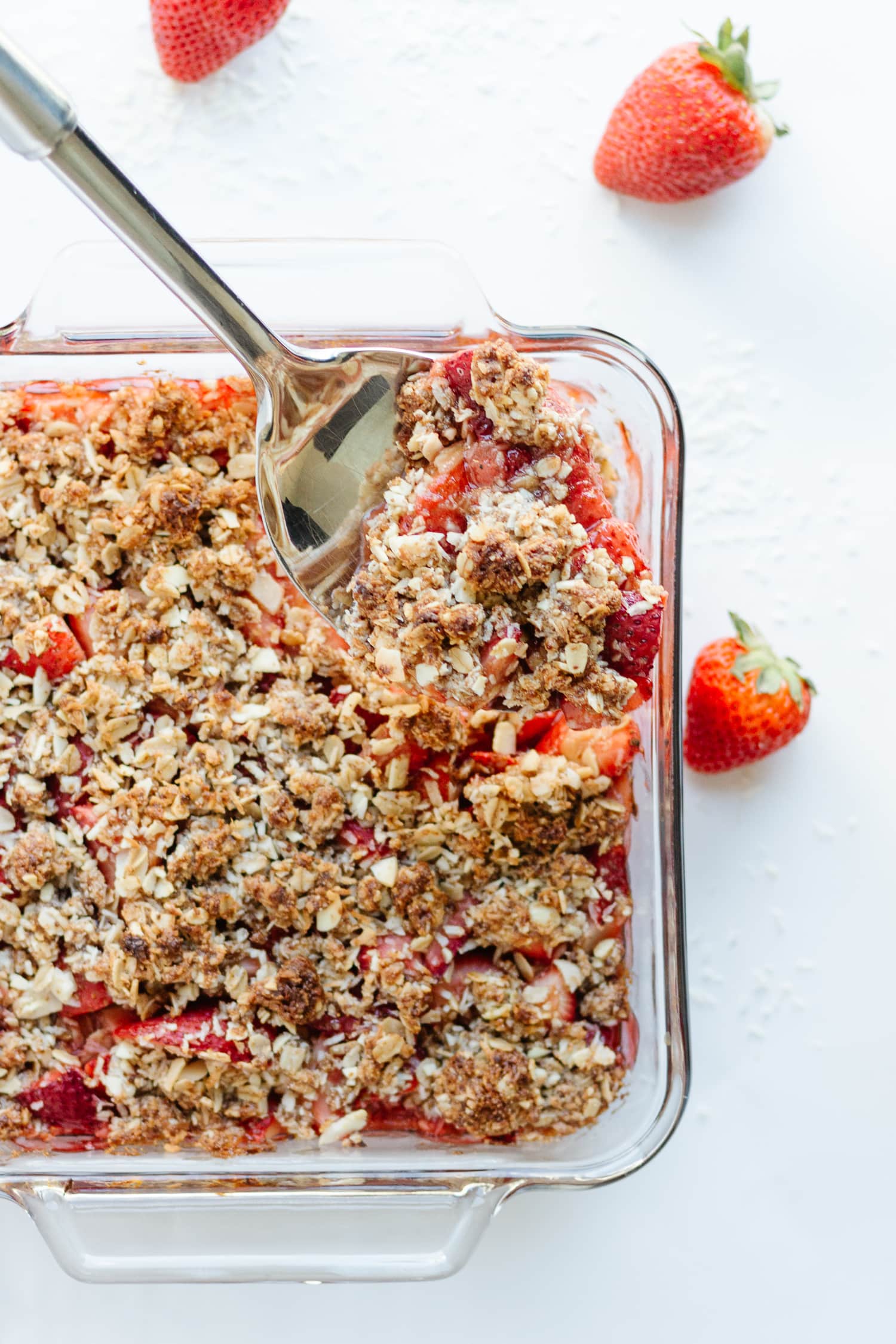 Square glass dish of strawberry coconut crisp with large metal serving spoon lifting out a portion.