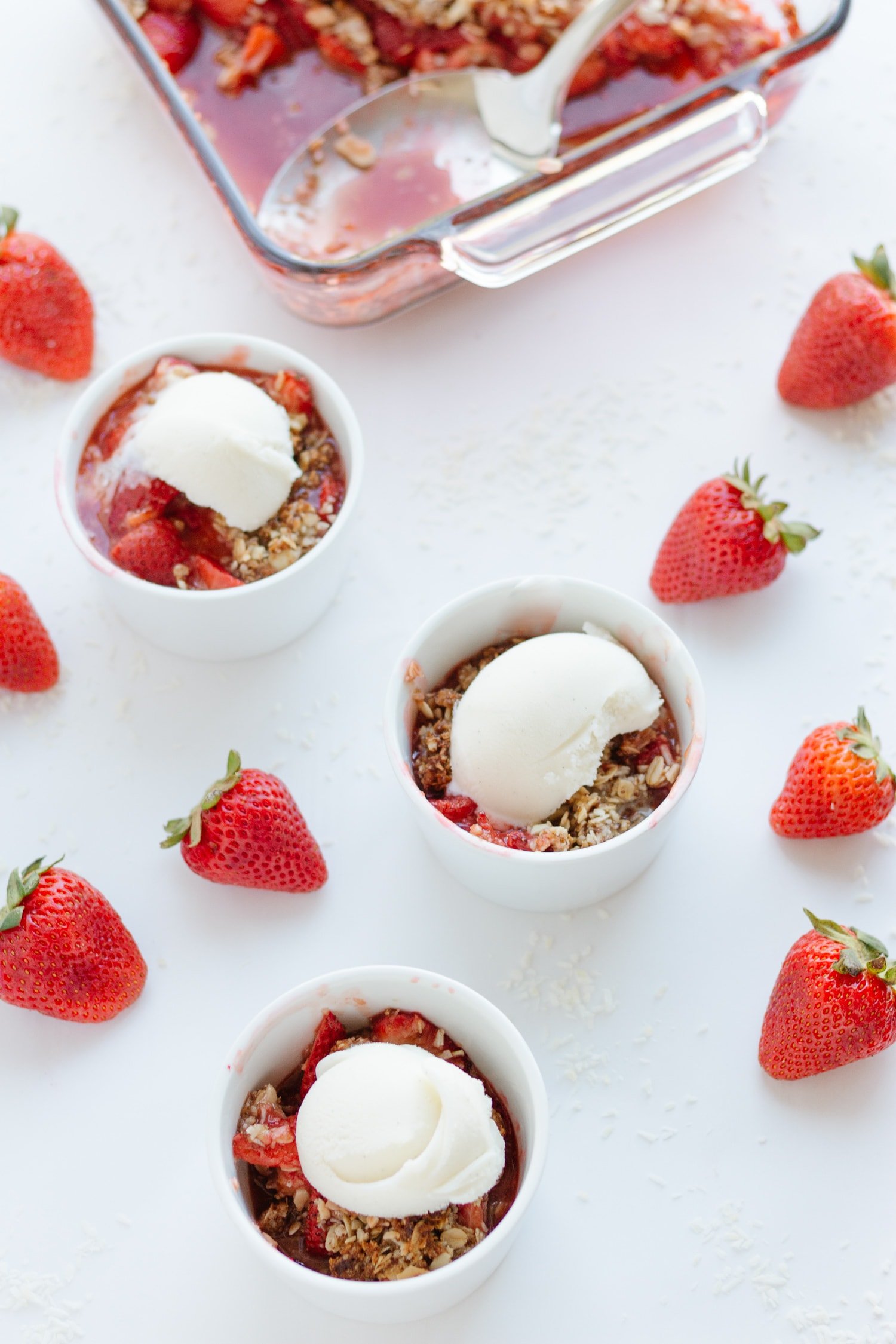 Three ramekins of strawberry coconut crisp topped with ice cream  with baking dish in background and strawberries scattered on white surface.