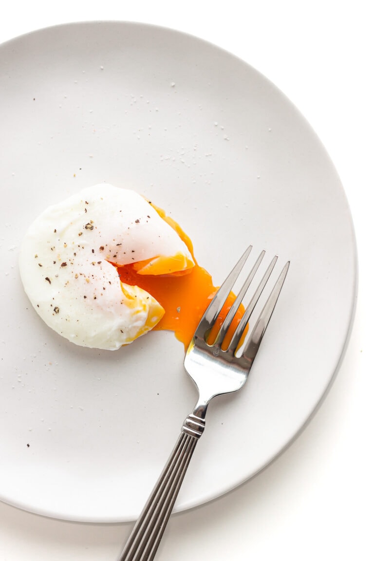 A single poached egg that has been cut into to release the oozy yolk on a white plate with fork.