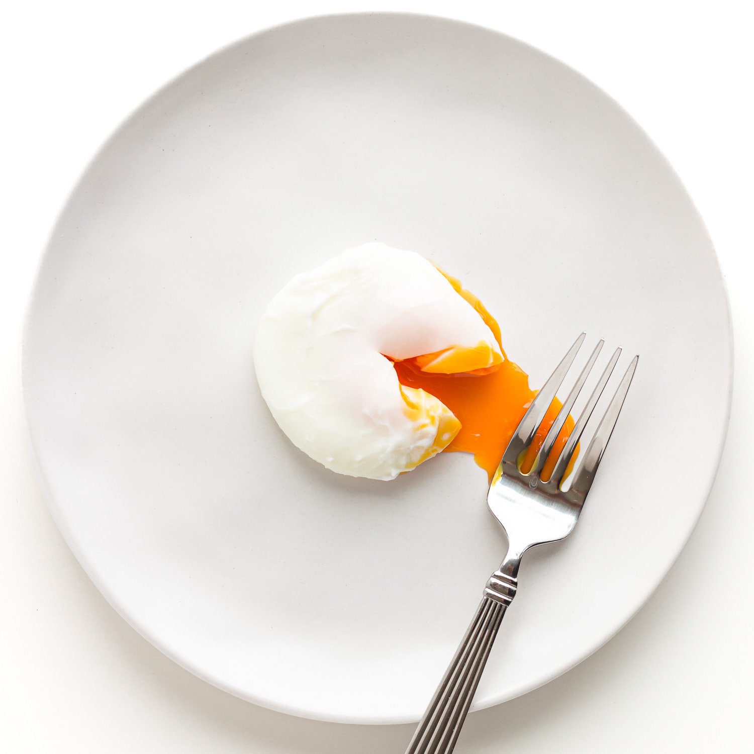 A poached egg on a white plate with a fork that has cut in to release some of the oozy yolk.