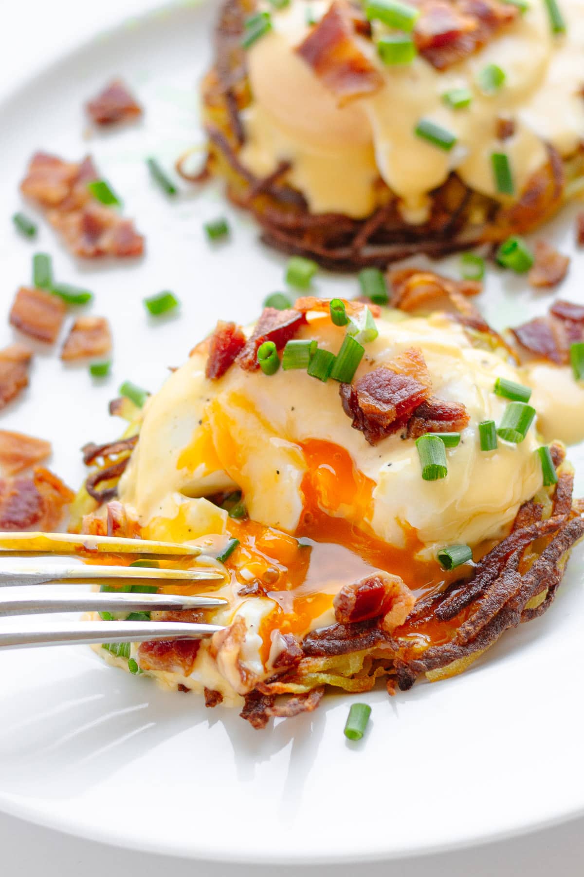 Potato rosti eggs Benedict garnished with chopped chives and crispy bacon pieces with a fork cutting into one.