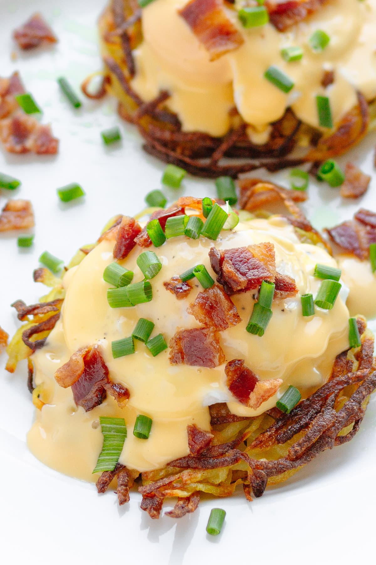 Two Rosti Eggs Benedict on a white plate garnished with chopped bacon and fresh chives.