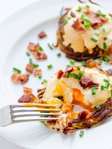 Two rosti eggs benny on a white plate topped with hollandaise sauce, bacon bits and chopped chives.