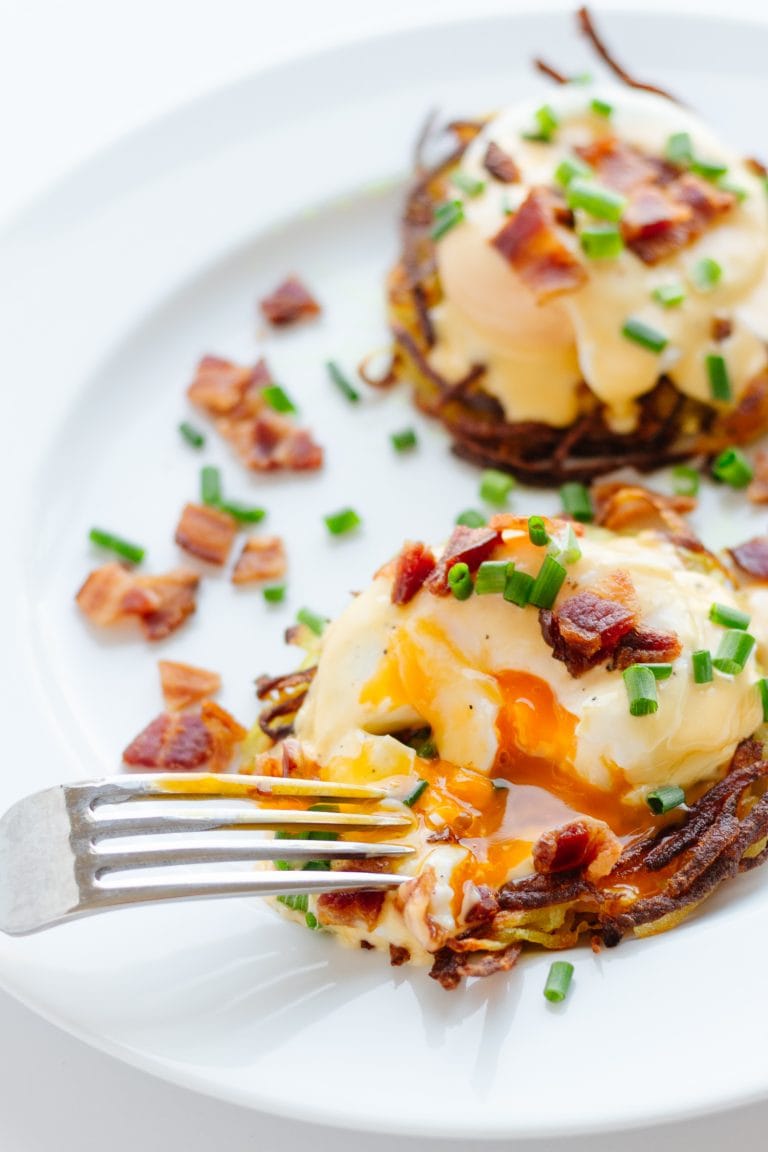 Two rosti eggs benny on a white plate topped with hollandaise sauce, bacon bits and chopped chives.