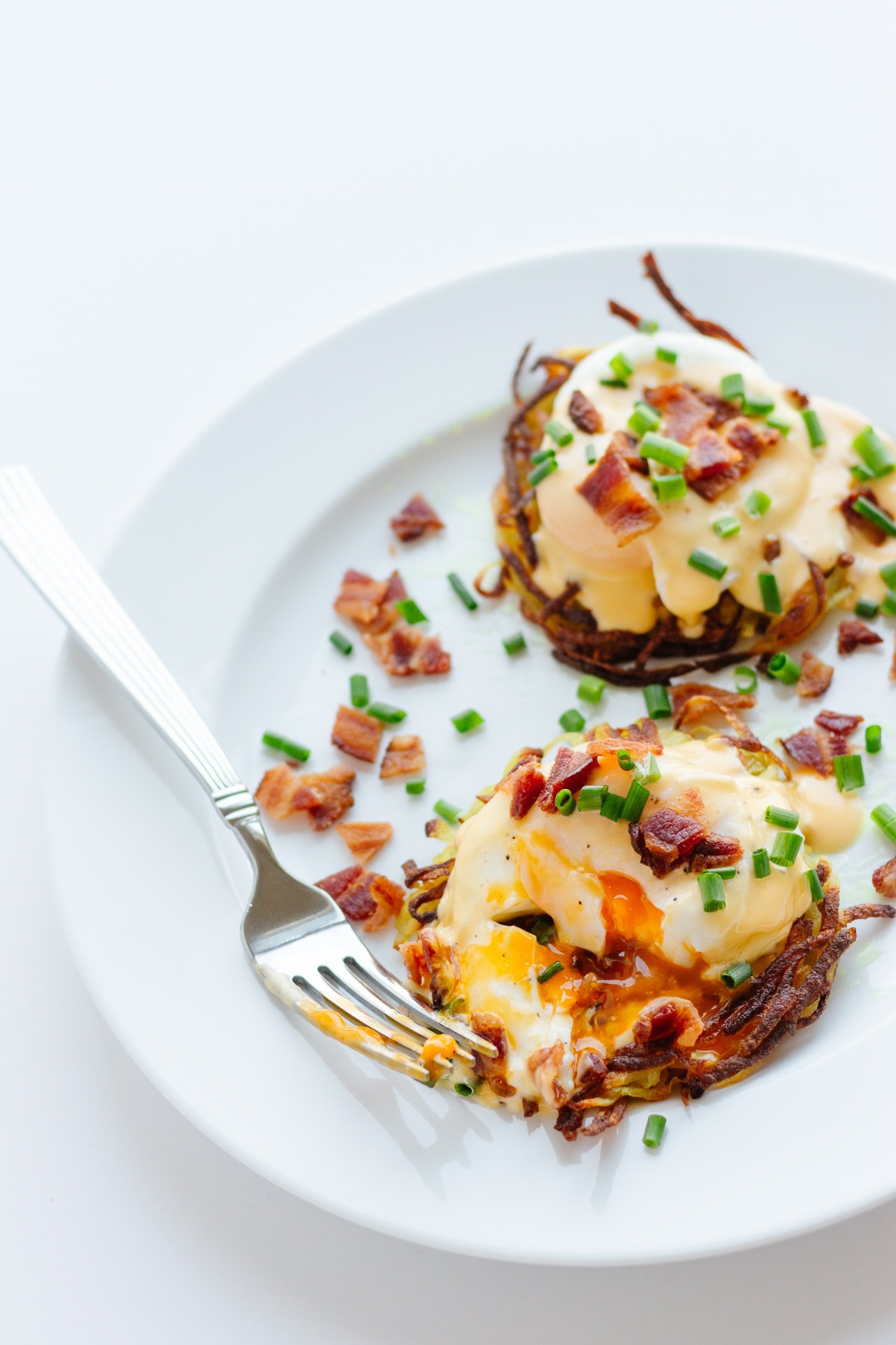 Two rosti eggs benny on a white plate with a yolk covered fork resting on the side