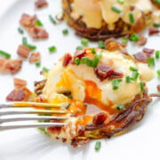 A fork cutting into Rosti Eggs Benny on a white plate garnished with chopped bacon and chives.