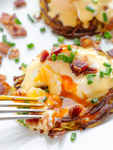 A fork cutting into Rosti Eggs Benny on a white plate garnished with chopped bacon and chives.