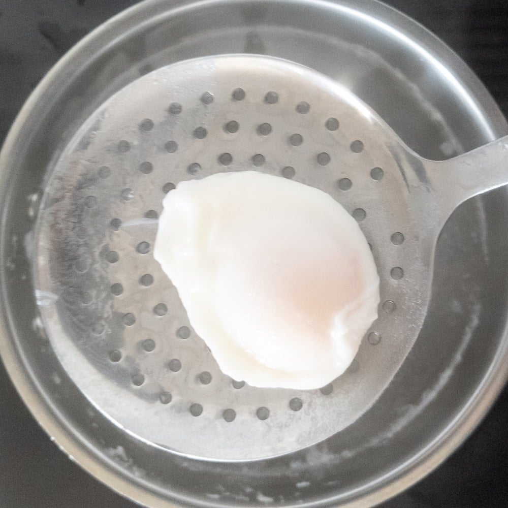 Pulling a poached egg out of saucepan with a stainless steel strainer spoon.
