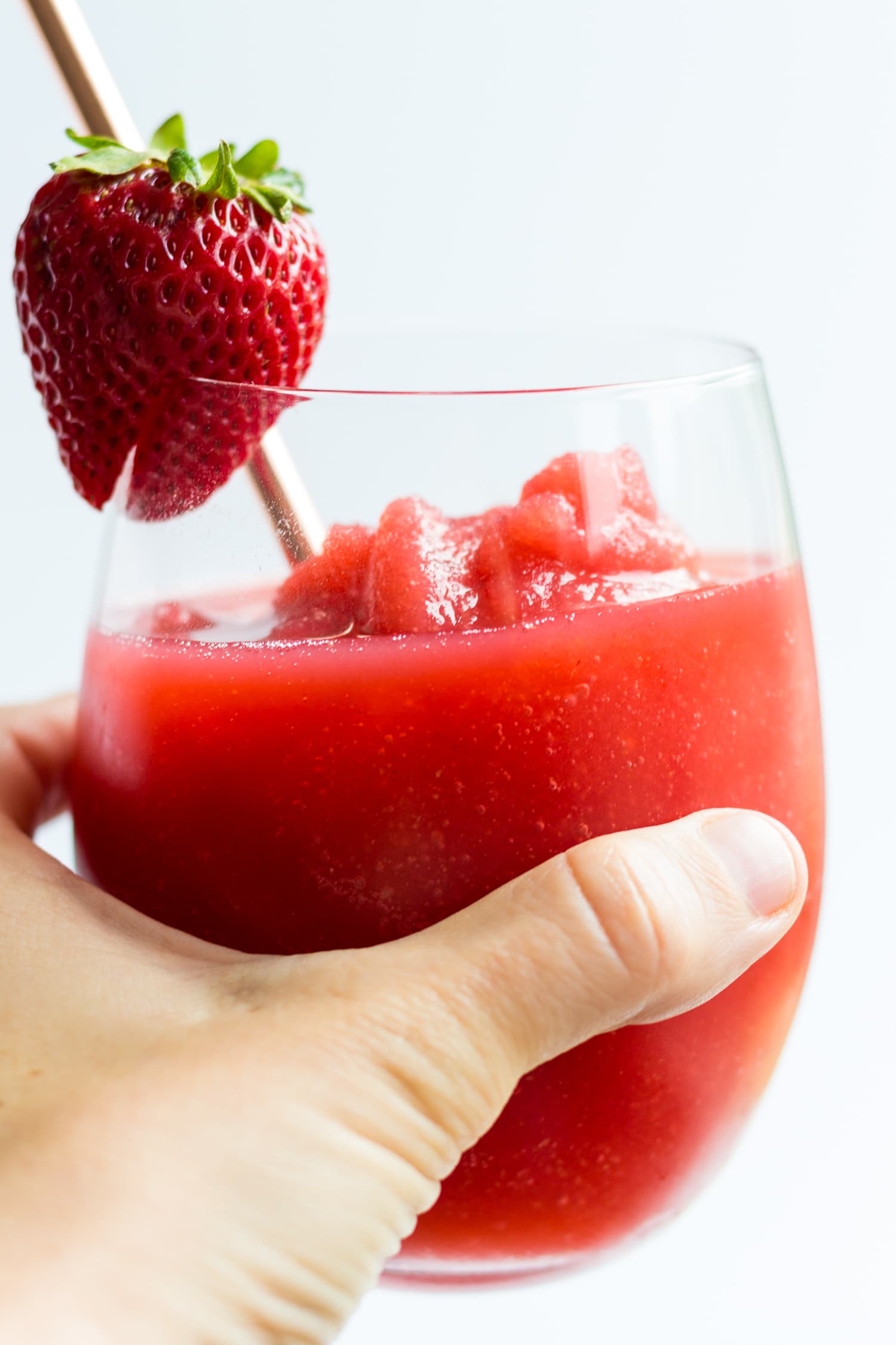 Hand holding a glass of strawberry frosé garnished with a fresh strawberry and rose gold straw