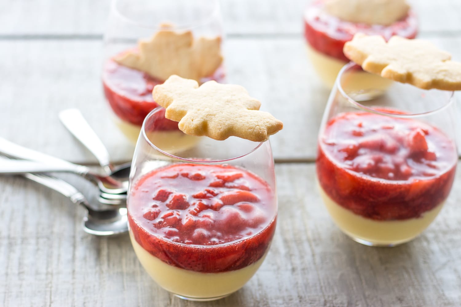 Four glasses of maple custard with strawberry compote that are topped with maple leaf shaped sugar cookies