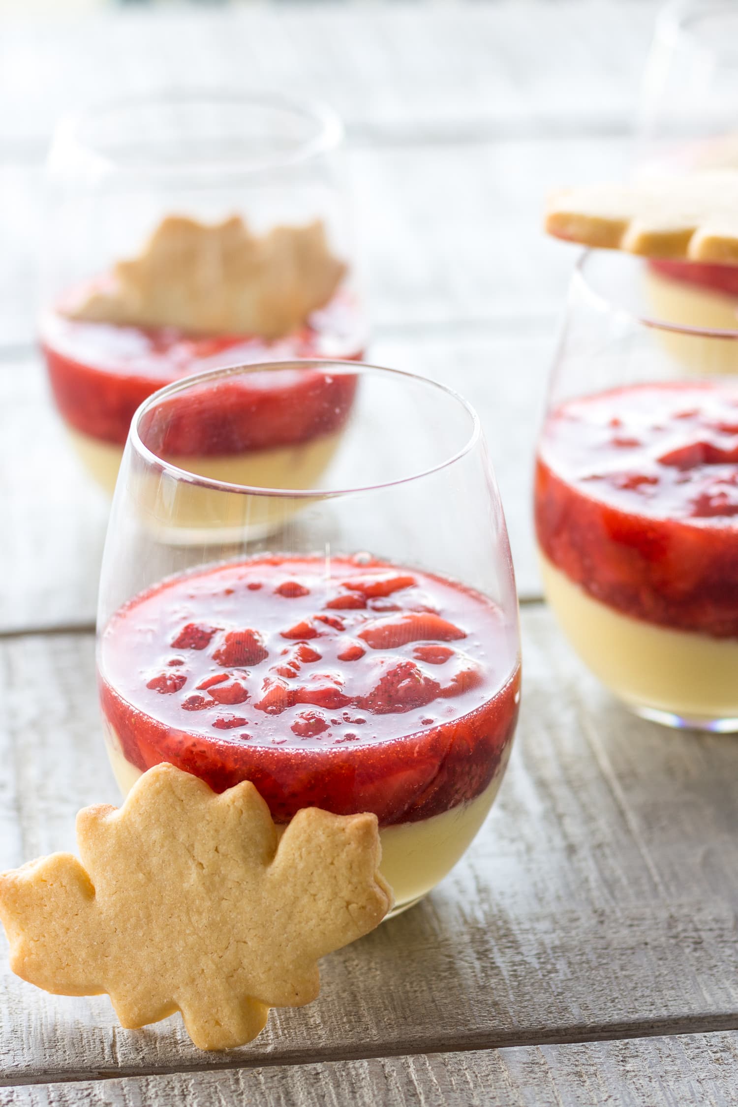 Three glasses of maple custard topped with strawberry compote and garnished with a maple leaf shaped sugar cookie.
