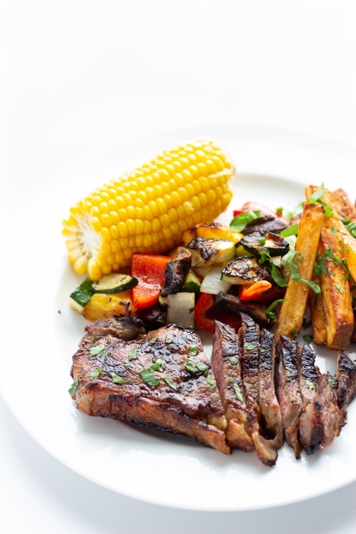 Grilled steak dinner on a white plate containing steak, corn on the cob, grilled mixed vegetables and roasted potatoes