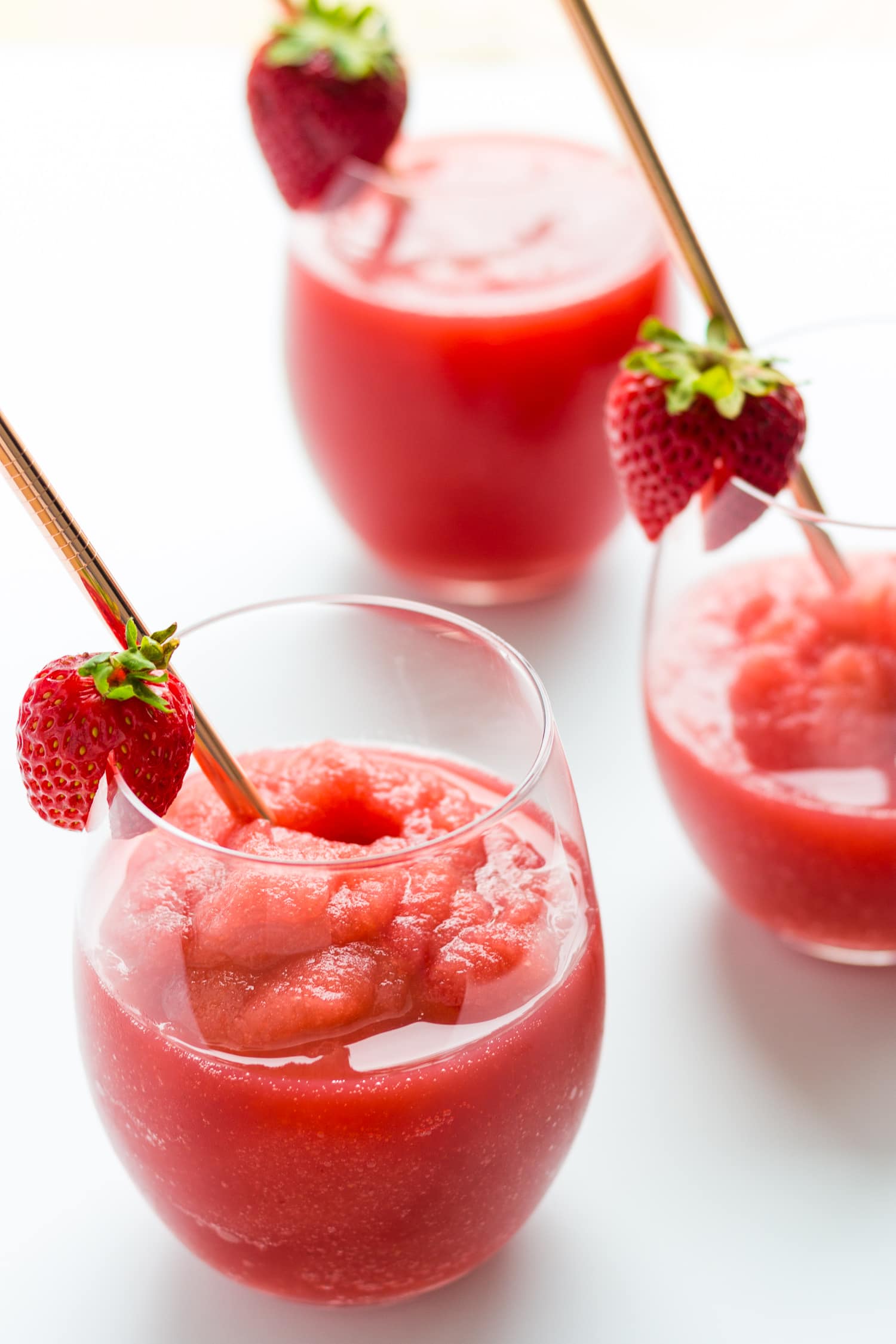 Three glasses of strawberry frosé garnished with a fresh strawberry and rose gold straw on a white surface