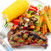 A grilled marinated steak on a white plate with some side dishes (corn, grilled veggies and potato wedges).