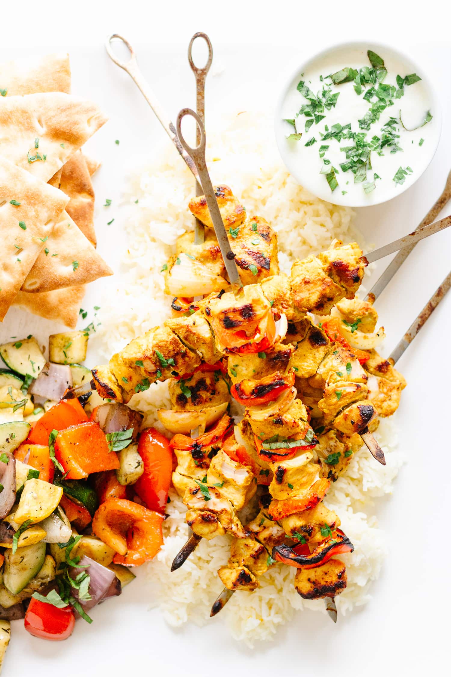 Grilled Tandoori Chicken Kebabs over a bed of rice with cucumber raita, naan bread and grilled veggies in the corners.
