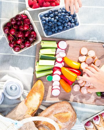 Overhead photo of a picnic set up on a blanket with two childrens hands reaching in.