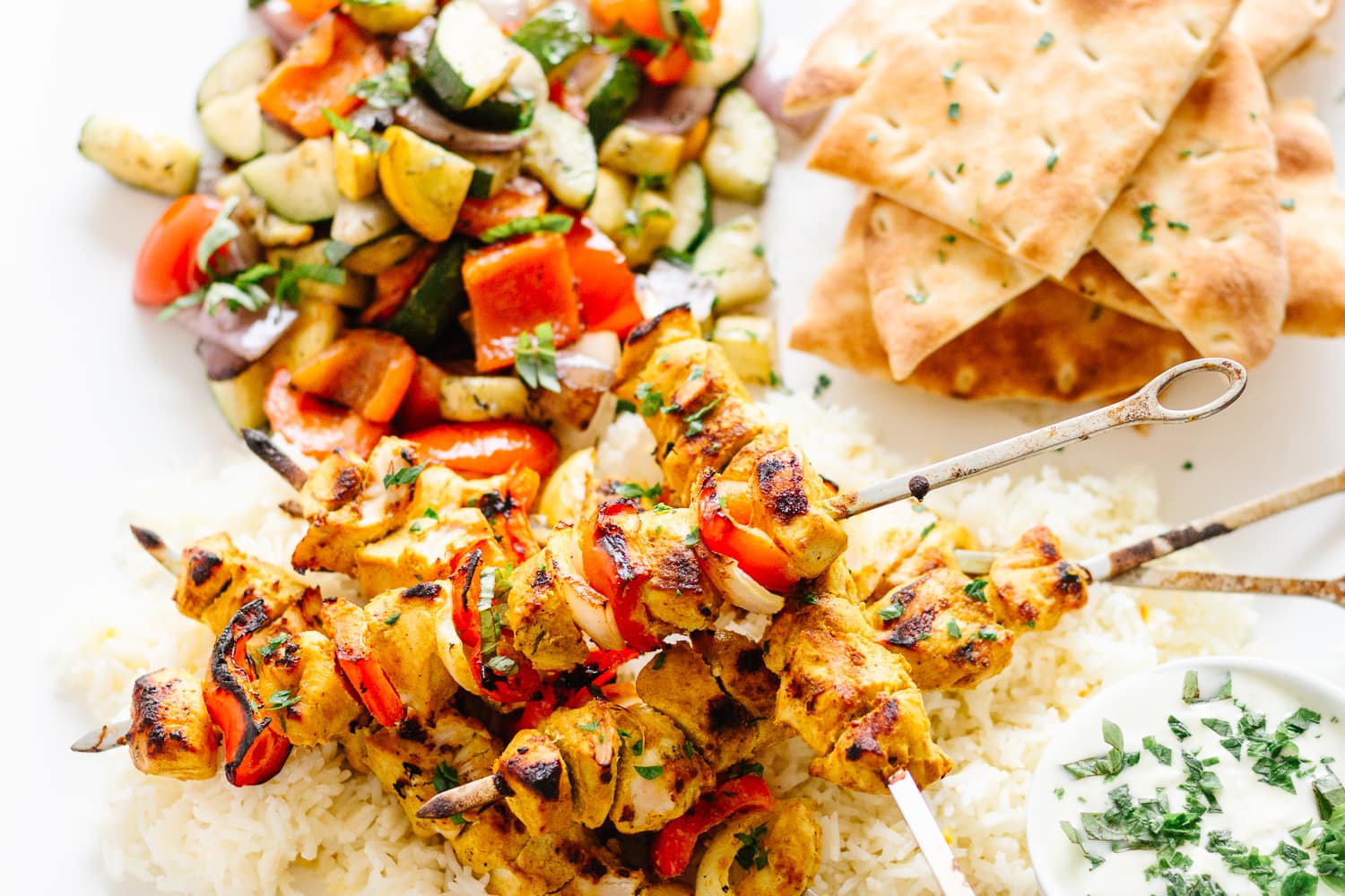 Platter containing grilled tandoori chicken kebabs on a bed or rice, naan bread, grilled veggies and small dish of cucumber raita.