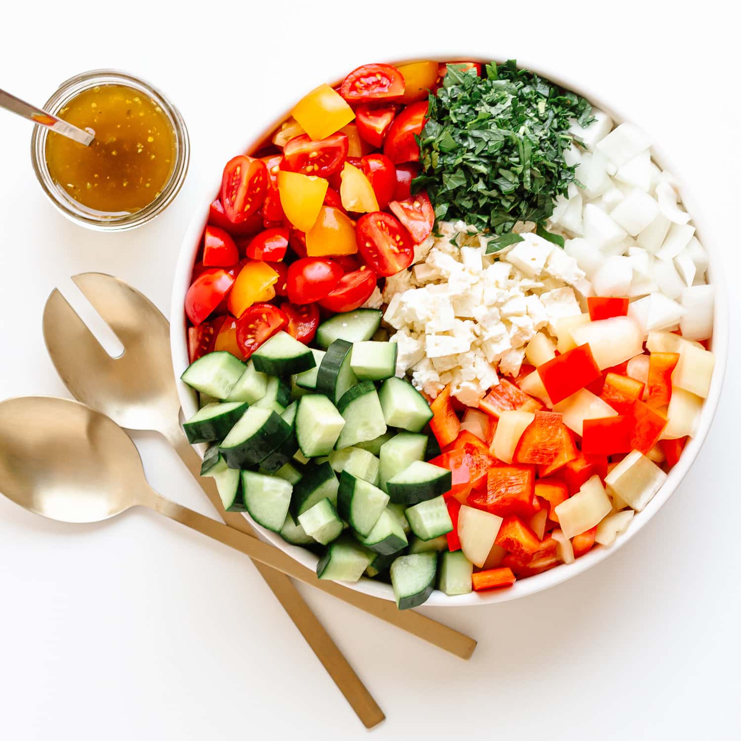 Variety of chopped veggies arranged separately in a bowl with dressing and salad servers on the side.