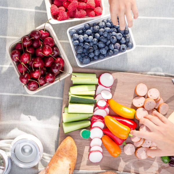 Picnic set up on a picnic blanket with two children's hands reaching in to grab food.