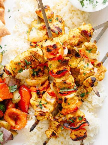 Grilled Tandoori Chicken Kebabs piled on some rice and surrounded by other sides.