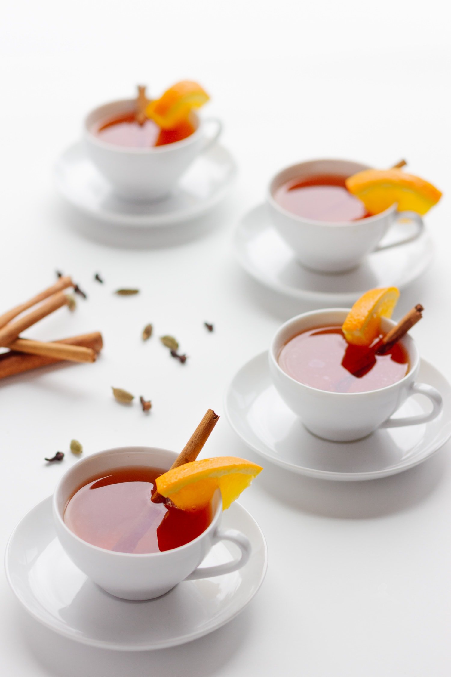 4 white tea cups arranged in a C pattern filled with Orange Spice Mulled Tea and decorated with an orange slice and cinnamon stick.