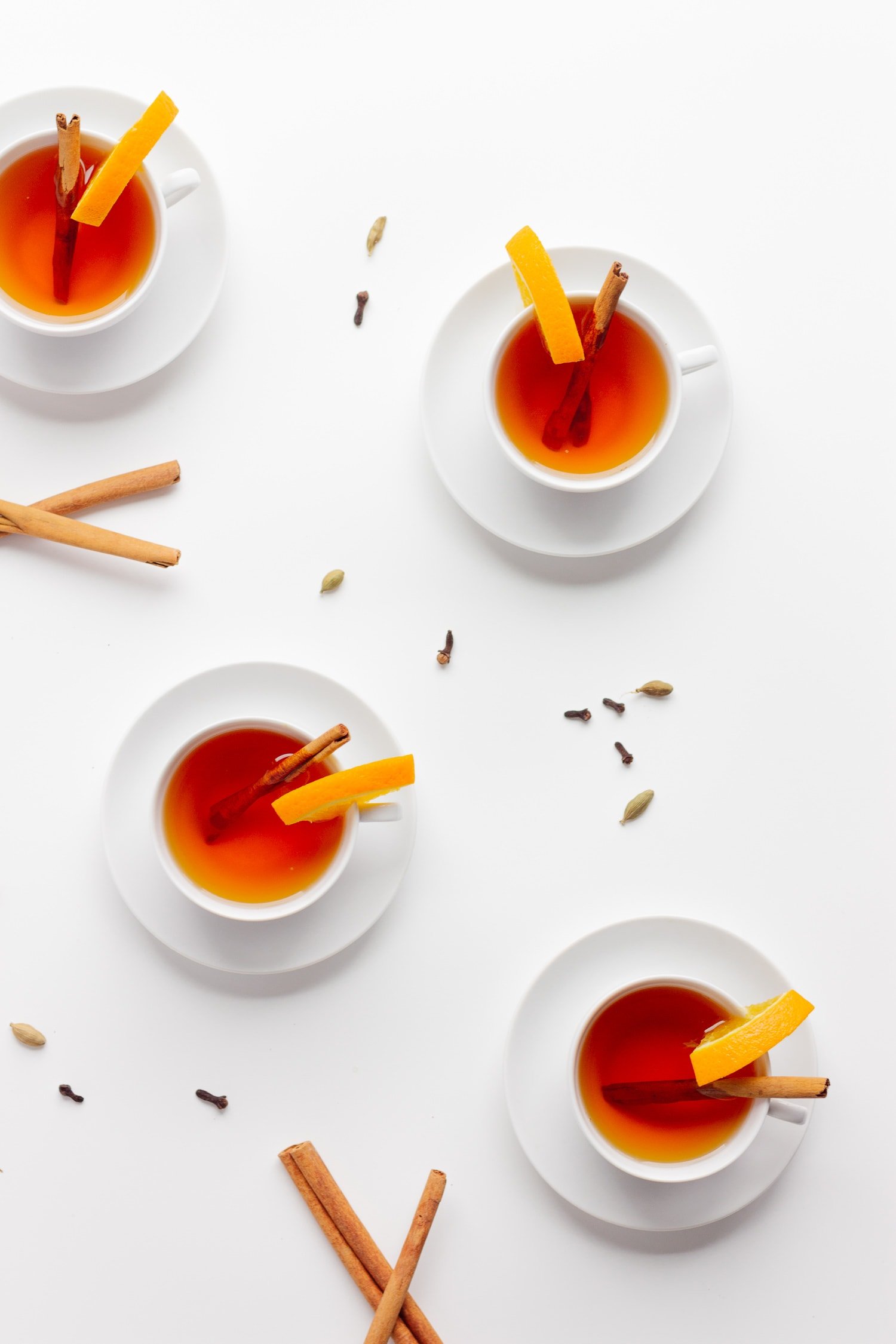 Overhead shot of 4 white tea cups filled with Orange Spice Mulled tea, decorated with an orange slice and cinnamon stick with cinnamon sticks, cloves and cardamom scattered around