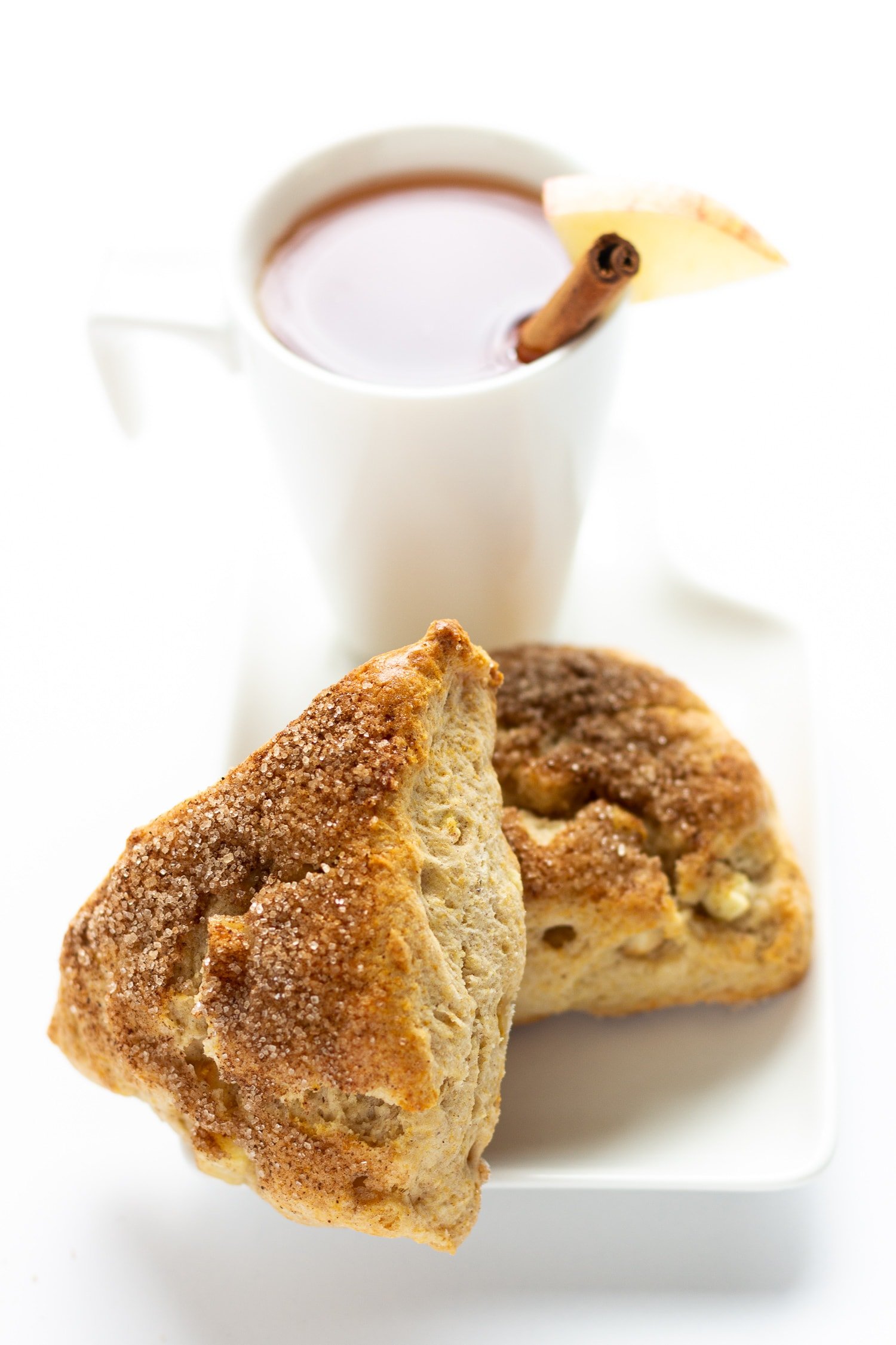 Two apple cinnamon scones on a white plate with a white mug of apple cider in the background