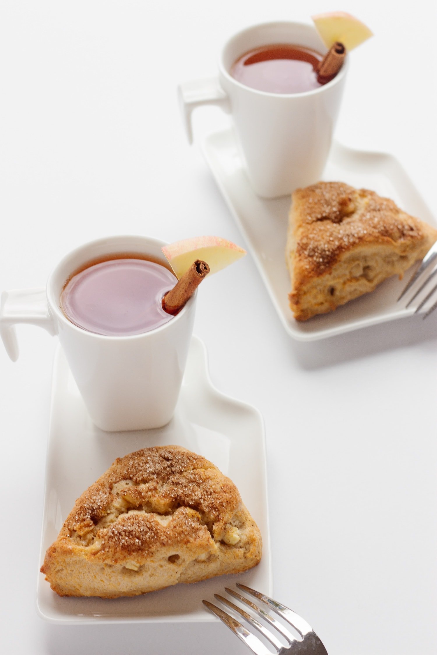 Two white dessert plates each containing an apple cinnamon scone and cup of hot apple cider.