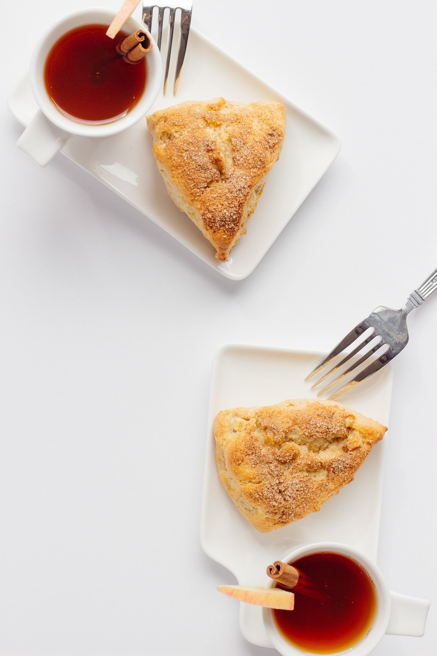 Two white dessert plates each holding an apple cinnamon scone and cup of hot apple cider with forks against the edge of the plate.