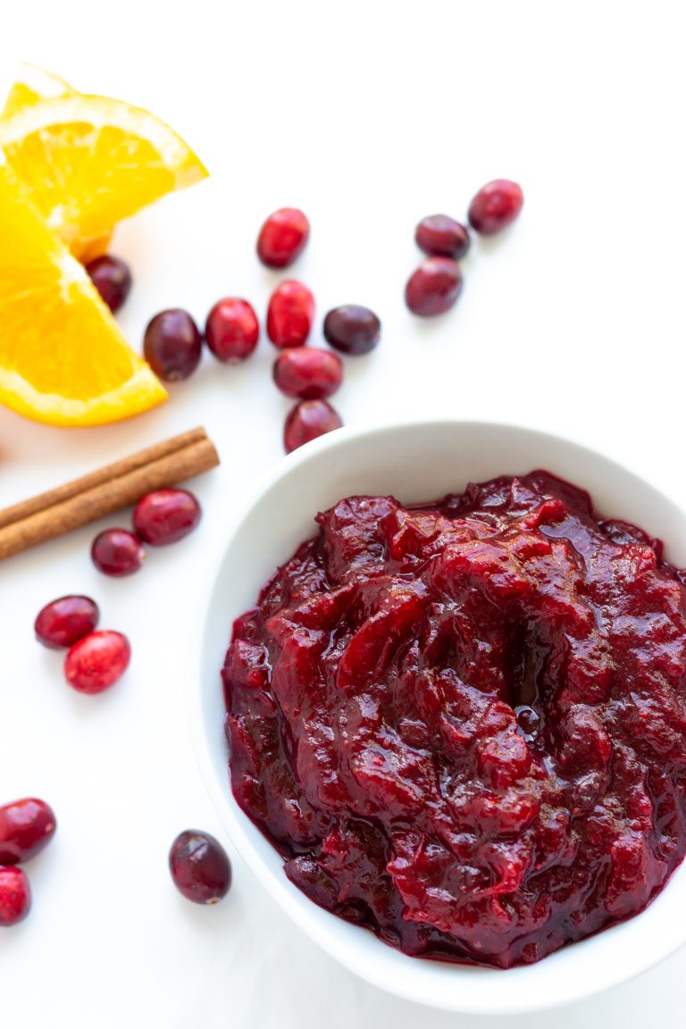 White bowl of cranberry orange sauce surrounded by cranberries, orange slices and cinnamon stick.