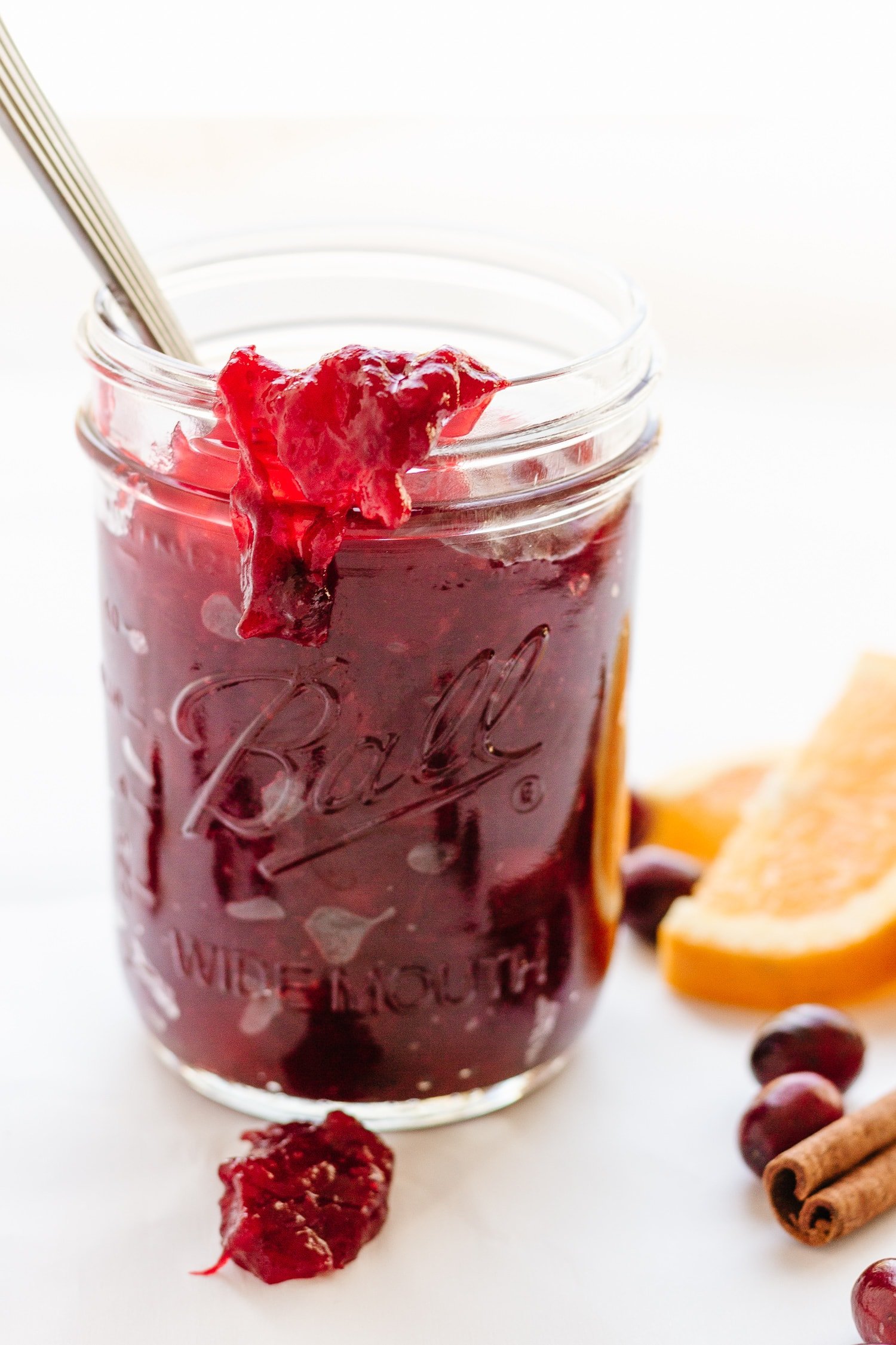 A mason jar filled with cranberry orange sauce and a spoon sticking out.  Orange slices, cranberries and a cinnamon stick are scattered next to the jar.