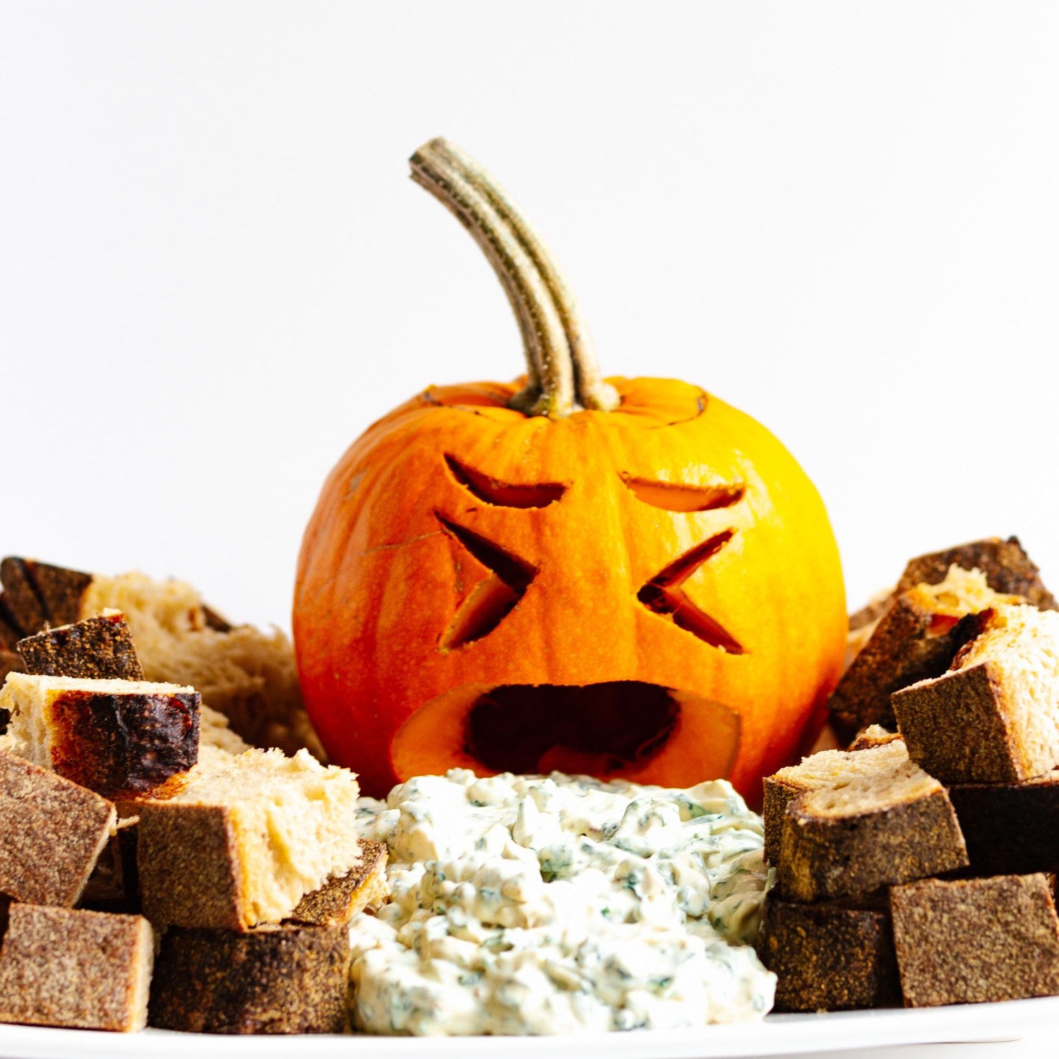 Small carved jack o' lantern with spinach dip pouring out of its mouth and surrounded by bread cubes.