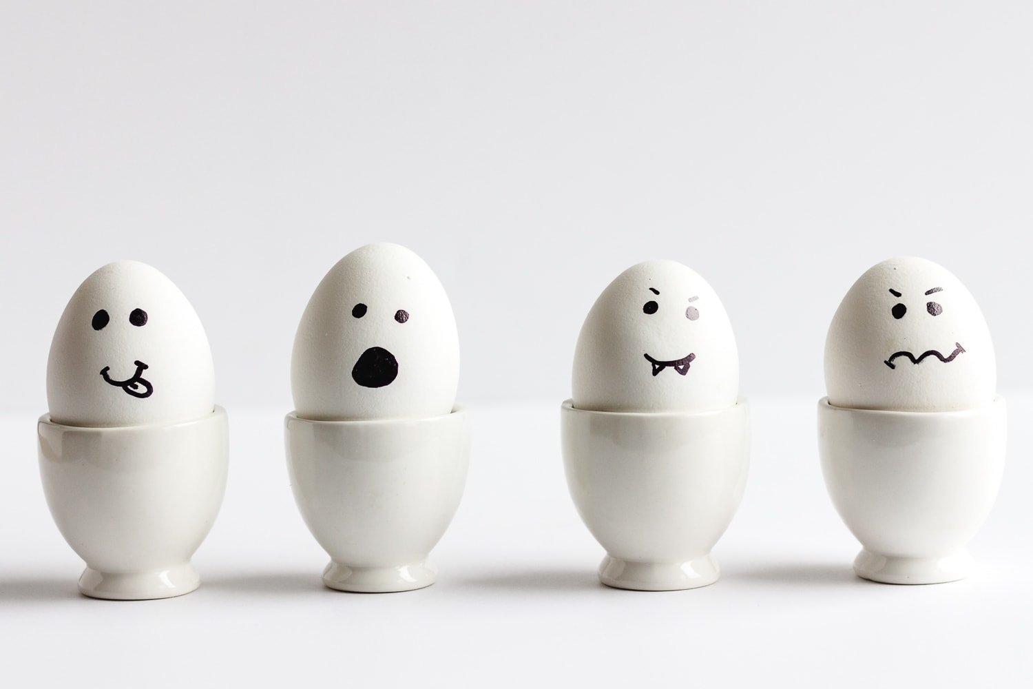 Four hardboiled eggs in white egg holders with faces drawn on to resemble Halloween ghosts.