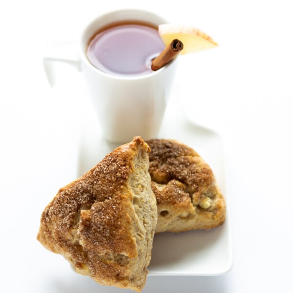 Two apple cinnamon scones with a white mug of hot apple cider sitting behind.
