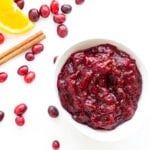 Cranberry Orange Sauce in a white bowl sitting on a white background with some fresh cranberries, orange slice and cinnamon stick scattered next to it.