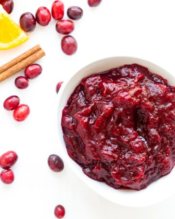 Cranberry Orange Sauce in a white bowl sitting on a white background with some fresh cranberries, orange slice and cinnamon stick scattered next to it.