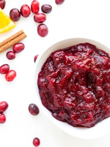 Cranberry Orange Sauce in a white bowl sitting on a white background with some fresh cranberries, orange slice and cinnamon stick scattered next to it.