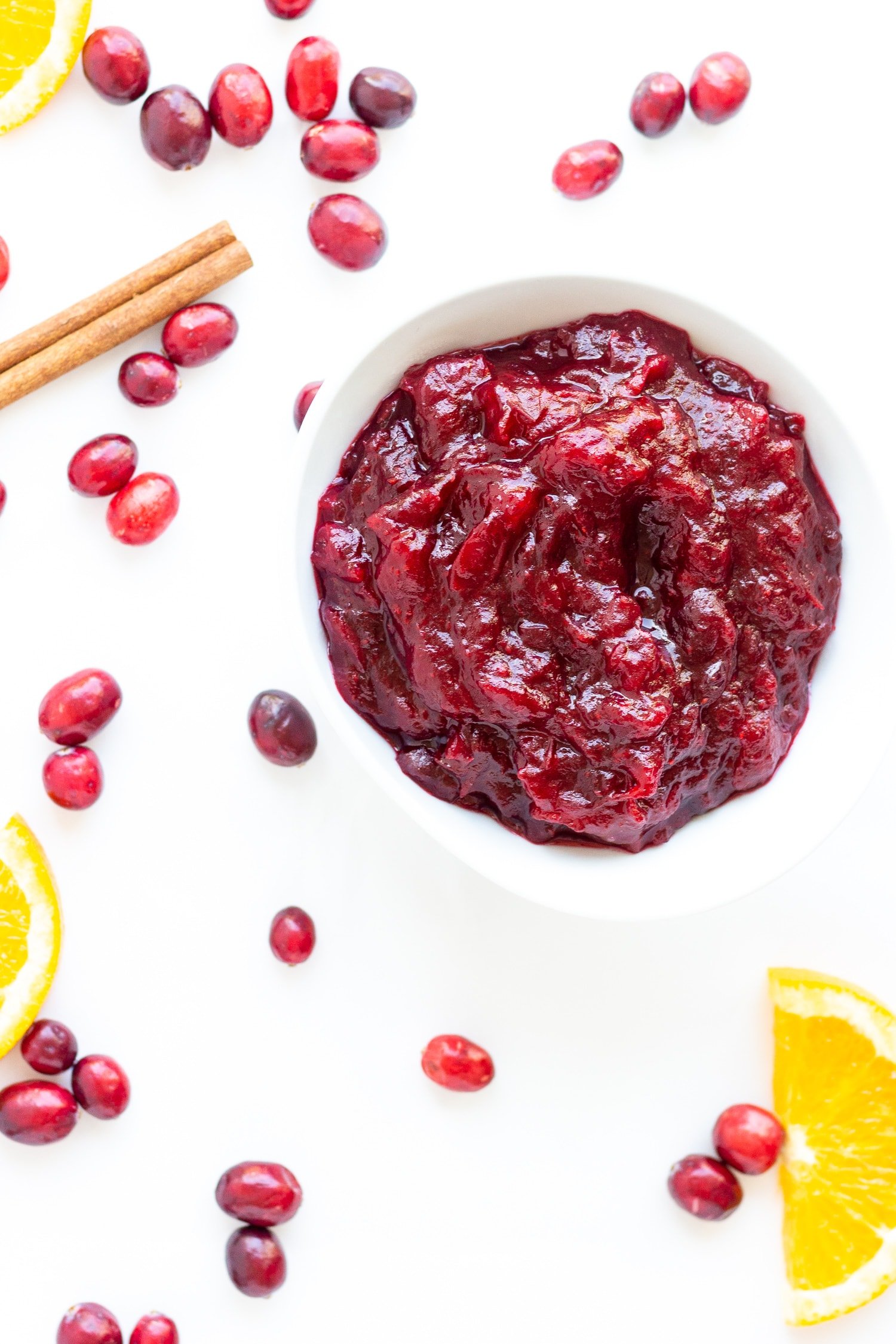 White bowl of cranberry sauce sitting on a white background with fresh cranberries, orange slices and a cinnamon stick scattered around it.