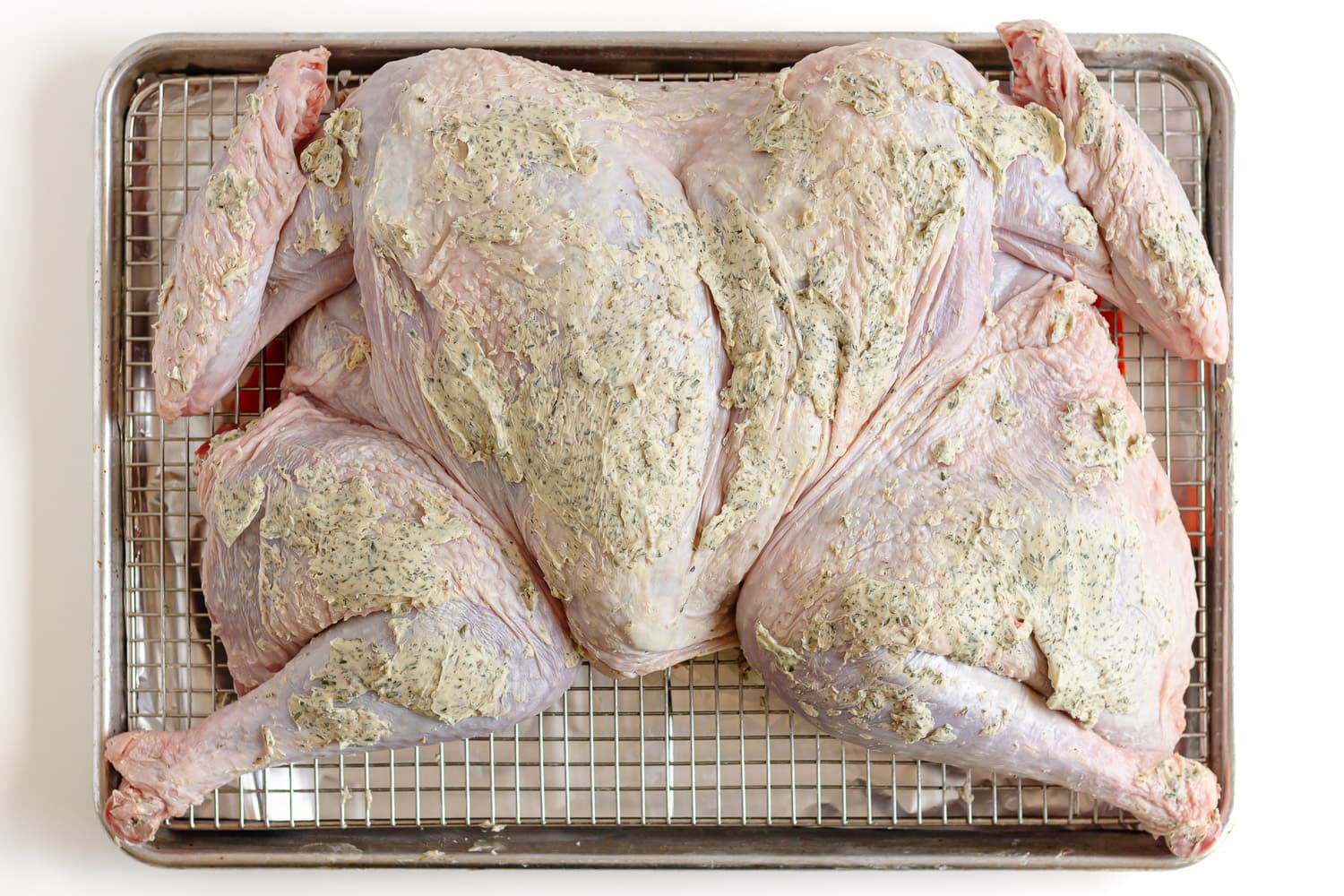 Herb butter slathered on a raw spatchcock turkey on a baking sheet.
