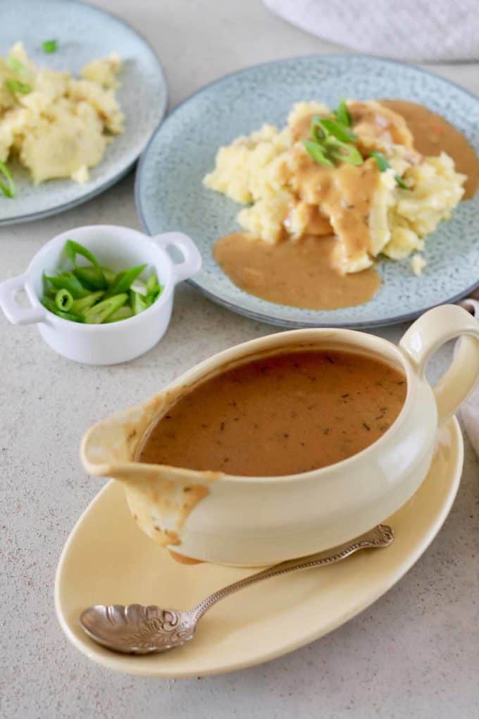 Vegan Gravy in a gravy boat with plates of mashed potatoes covered in gravy and garnished with green onion slices in the background