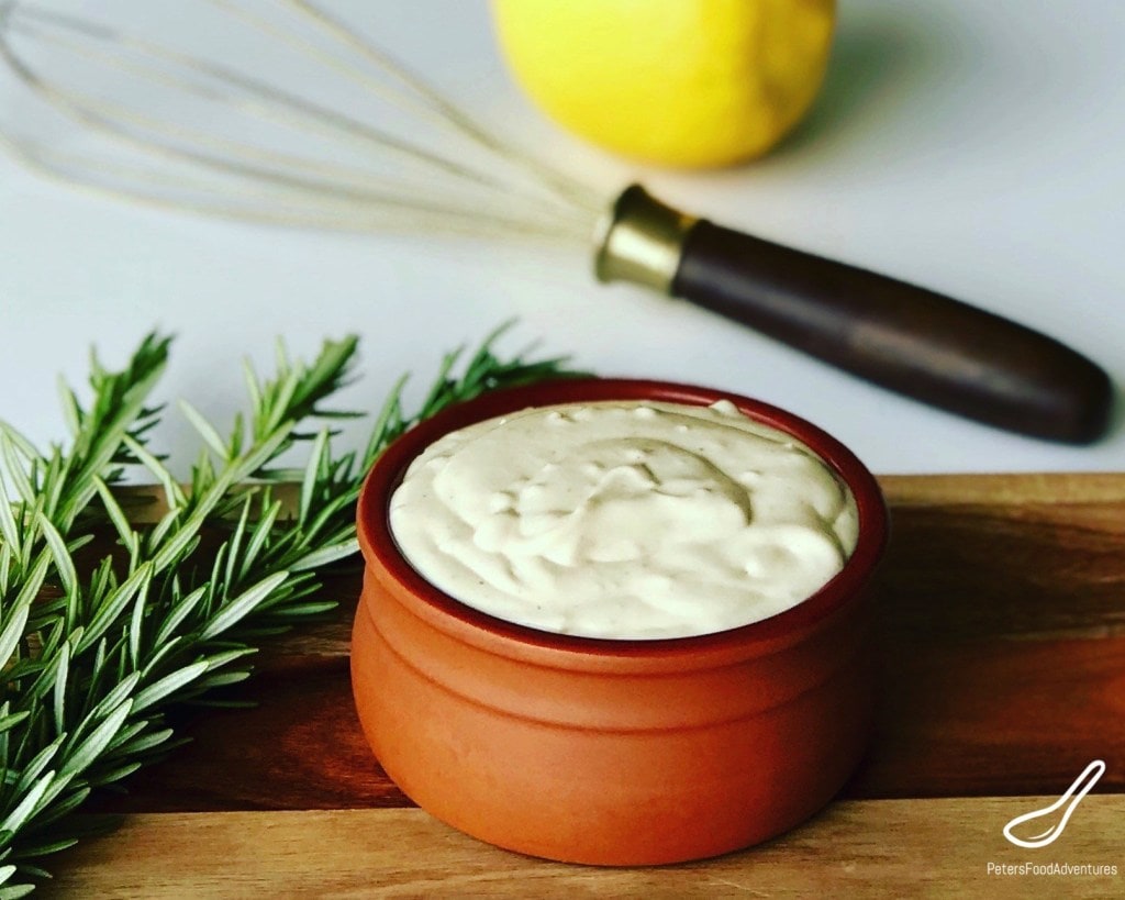 Creamy horseradish sauce in a bowl set on a wooden cutting board with sprig of rosemary next to it.  Whisk and lemon in the background