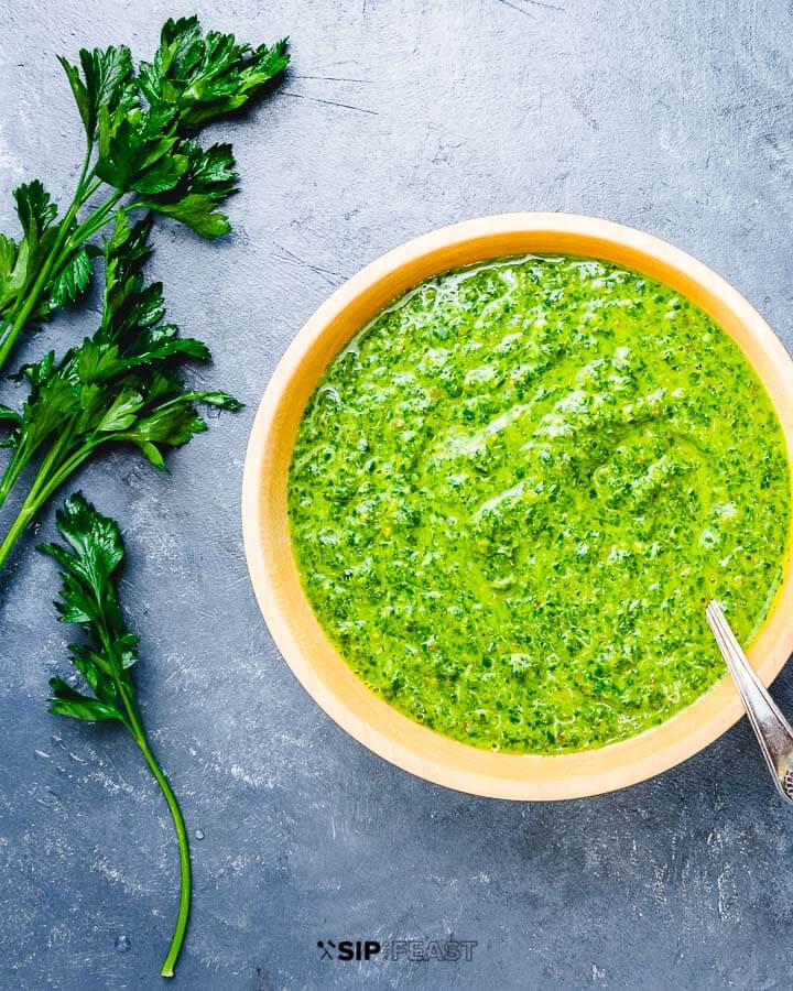 Italian Salsa Verde in an orange bowl with spoon sticking out.  Set on a concrete background with sprigs of Italian parsley.